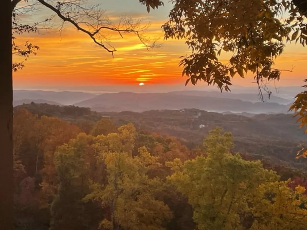 A breathtaking view capturing the sun setting behind rolling mountain ranges and autumnal trees. The warm, tranquil light invites quiet reflection and underscores the natural beauty of the location.