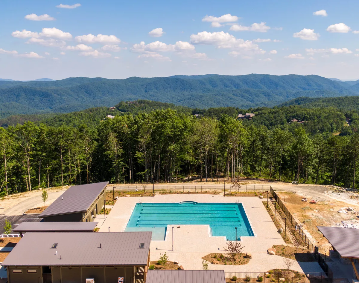 A modern swimming pool set amidst trees and mountains, with a tranquil blue surface reflecting the surrounding scenery. The pool area, bordered by stylish buildings and greenery, invites guests to swim, sunbathe, or simply relax in a serene outdoor setting.