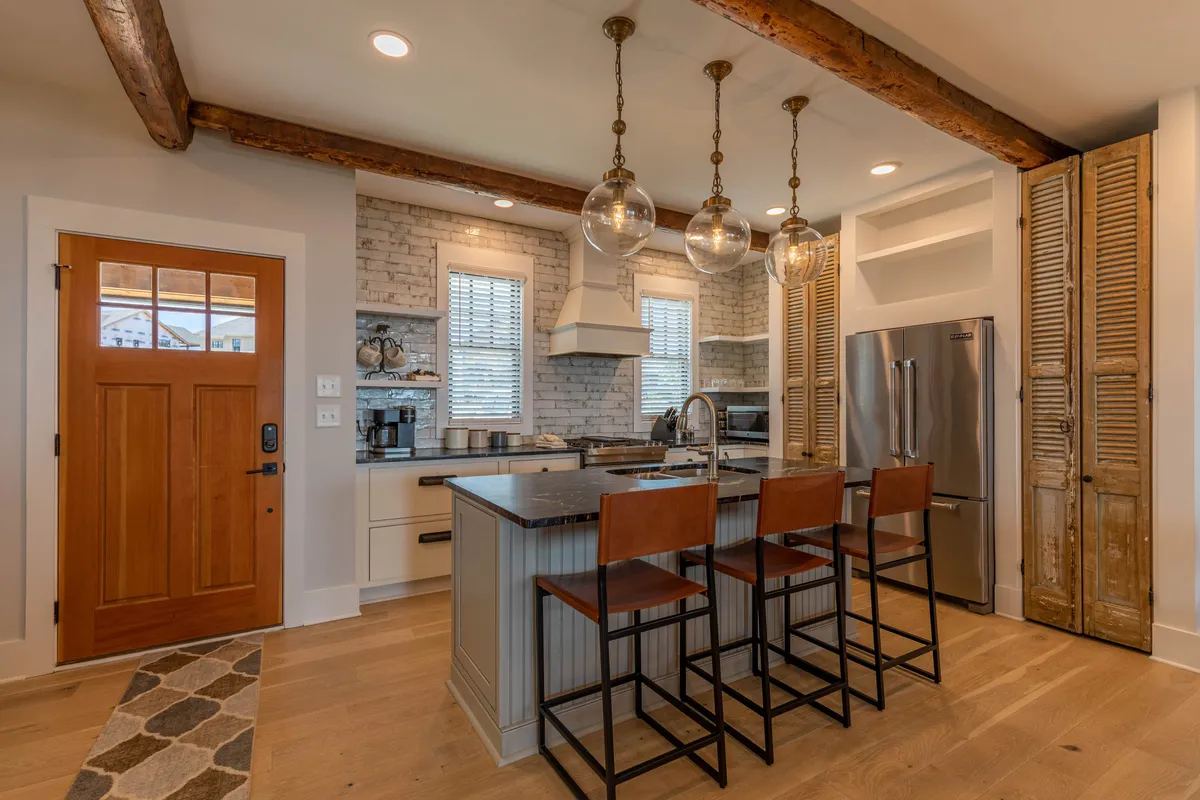 A warm, inviting kitchen space featuring a large kitchen island with hanging pendant lights, exposed wooden beams, and a clear view of the adjoining areas. The scene conveys an atmosphere perfect for cooking, entertaining, and family gatherings, with custom cabinetry and a sense of open flow.
