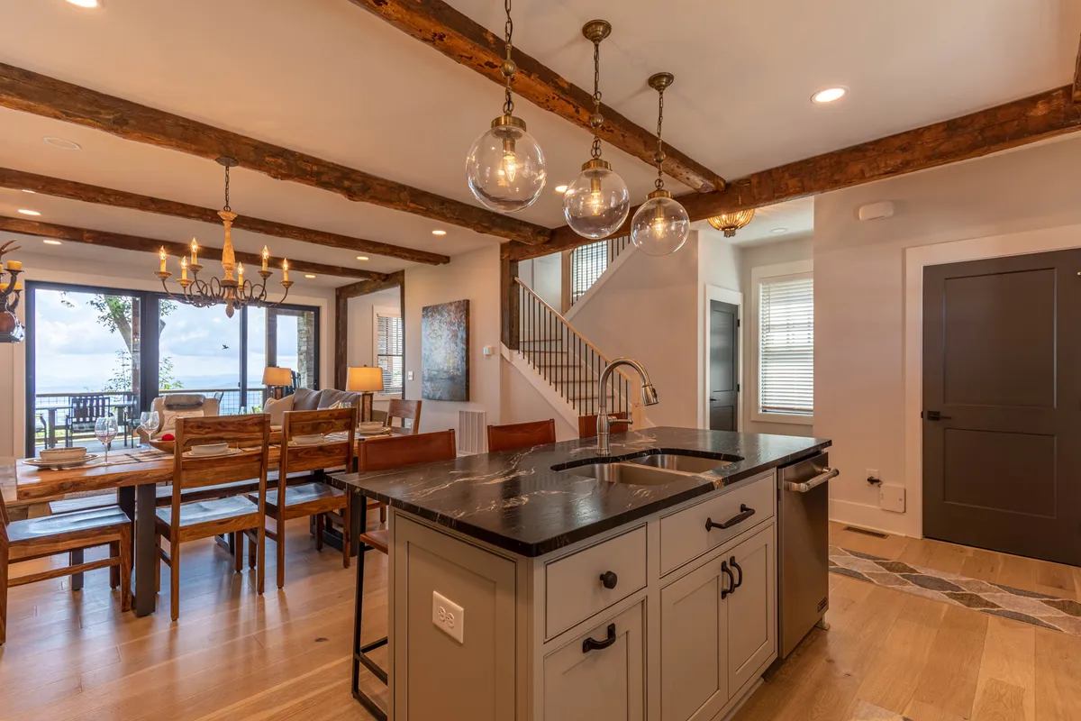 A bright and open area seamlessly integrating the kitchen and dining space. Features include a kitchen island, a sizable elegant dining table, large windows offering beautiful views, and chandeliers that add a sophisticated touch. The ambiance encourages gathering for meals and enjoying time together.