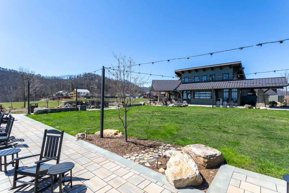 A welcoming exterior view featuring a wide lawn area, the main building structure, rock formations, and inviting outdoor seating. The green space stretches out, bordered by trees and natural landscaping, offering plenty of room for dogs to play or guests to relax outside.