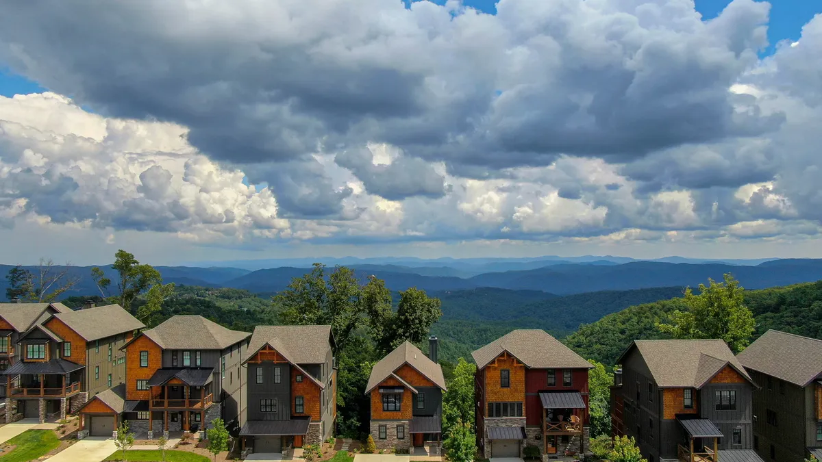 A serene mountain landscape with a row of houses nestled in the foreground, dramatic clouds overhead, and open spaces ideal for walks and relaxation. The view suggests accessibility to trails and the tranquil beauty of the Blue Ridge area.