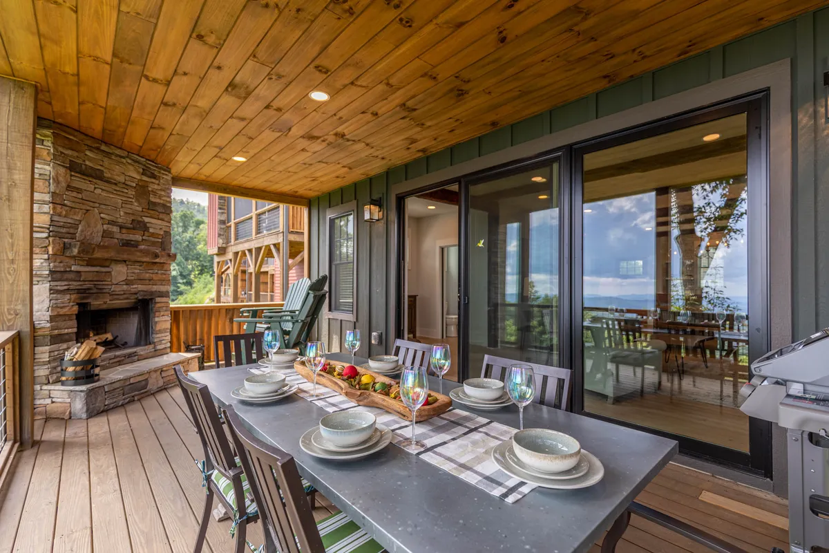 This exterior image showcases a covered deck furnished with a dining table and a built-in stone fireplace. The view through the glass doors extends to the surrounding mountain landscape, while the warm lighting and comfortable furnishings create an inviting setting for outdoor relaxation or dining.