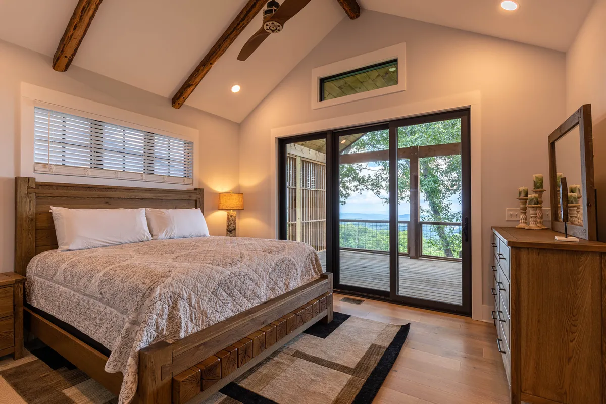 A serene bedroom featuring a wooden bed, large windows with a view of an outdoor deck, and a decorative mirror. The room exudes calmness and is designed for both relaxation and enjoying the scenery, with natural light streaming in.