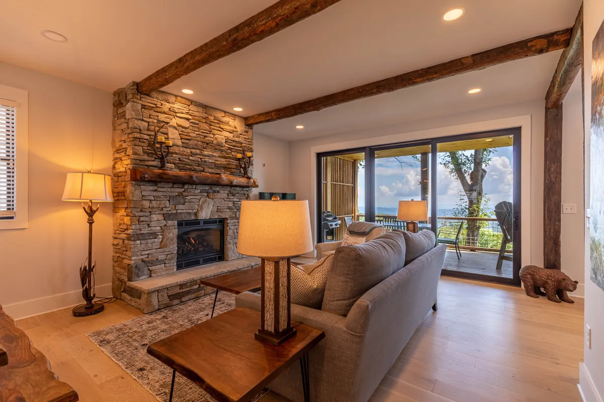 The image captures a warm, inviting living area centered around another striking stone fireplace. A comfortable sofa is perfectly positioned to enjoy the flames and look out through large windows at the mountain scenery. The room is filled with natural light, and the layout encourages both solitude and socializing, echoing the section’s emphasis on unwinding and togetherness.