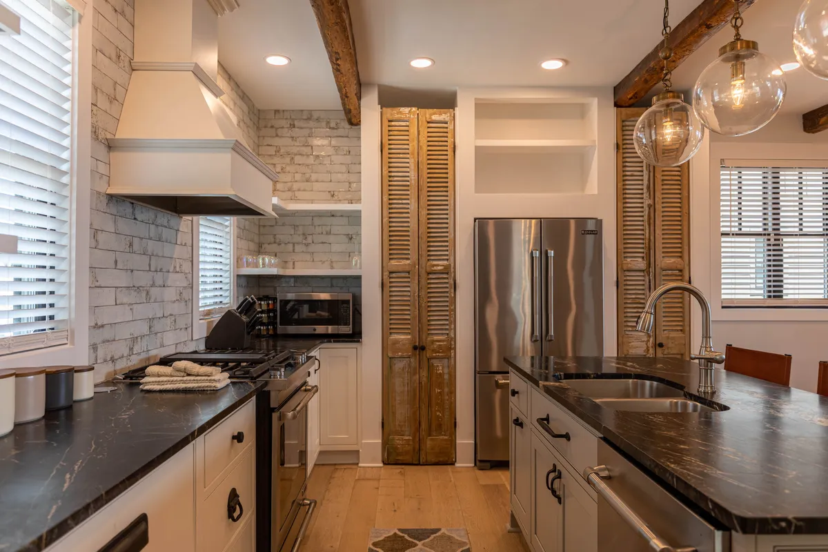 A modern kitchen with a prominent island, stainless steel appliances, wooden beams above, and an exposed brick wall providing a cozy, stylish backdrop. The layout showcases both functional cooking space and high-end finishes like granite counters and custom cabinetry.