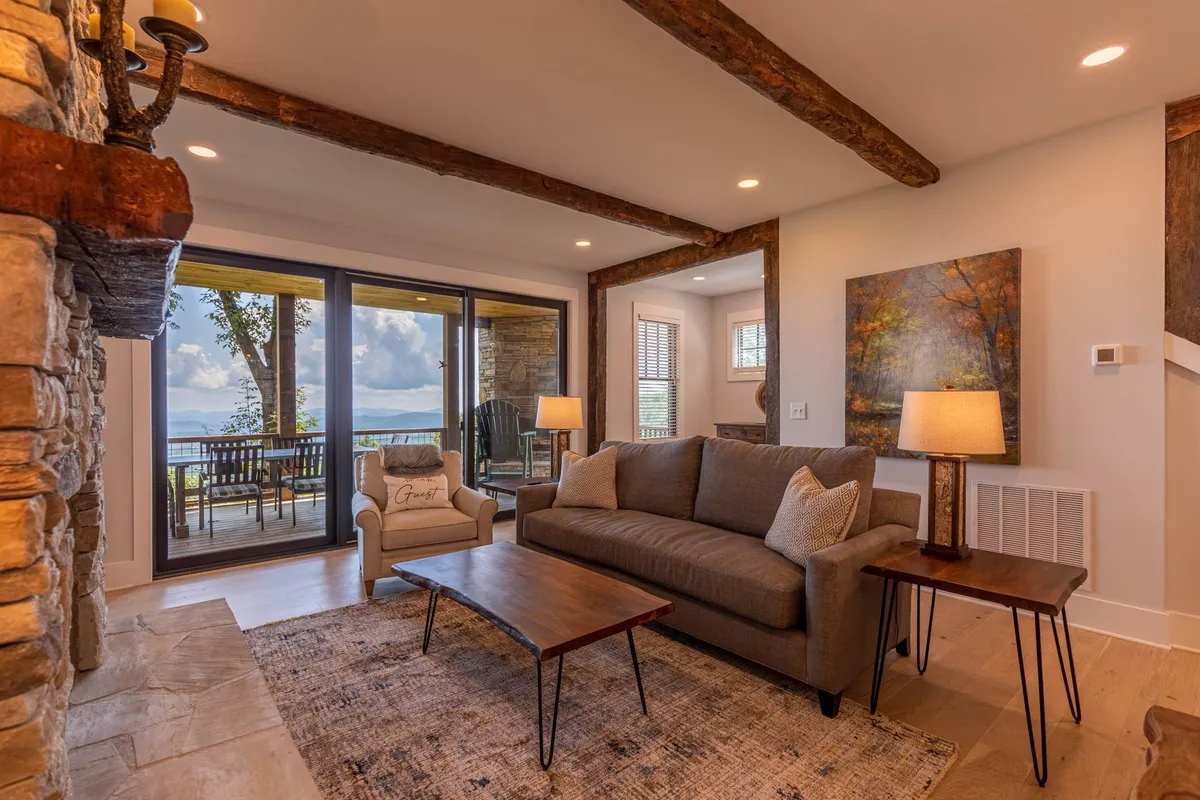 This image showcases a cozy living room with a prominent stone fireplace as the focal point. A plush sofa and coffee table are arranged near large windows, which offer sweeping views of the Blue Ridge Mountains. Artwork on the wall and accents of warm wood create a tranquil, inviting atmosphere. Natural light pours in, illuminating the space and highlighting the seamless connection between indoors and the surrounding landscape.